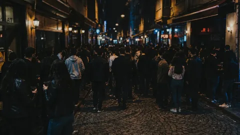 A dark, cinematic night photograph of a crowded bar street in Kadıköy, İstanbul, seen from behind the crowd. Dozens of people stand with their backs to the camera, drinks in hand, facing down a narrow wet-cobblestone street that recedes into deep shadow. Bars and pubs line both sides under warm wall lamps; a red neon PUB sign glows on the right, with cooler blue light further down the street. No faces are visible; everyone is turned away, dissolving into the movement of the street. In the lower-left foreground, two figures in leather jackets stand half-turned, holding plastic cups.